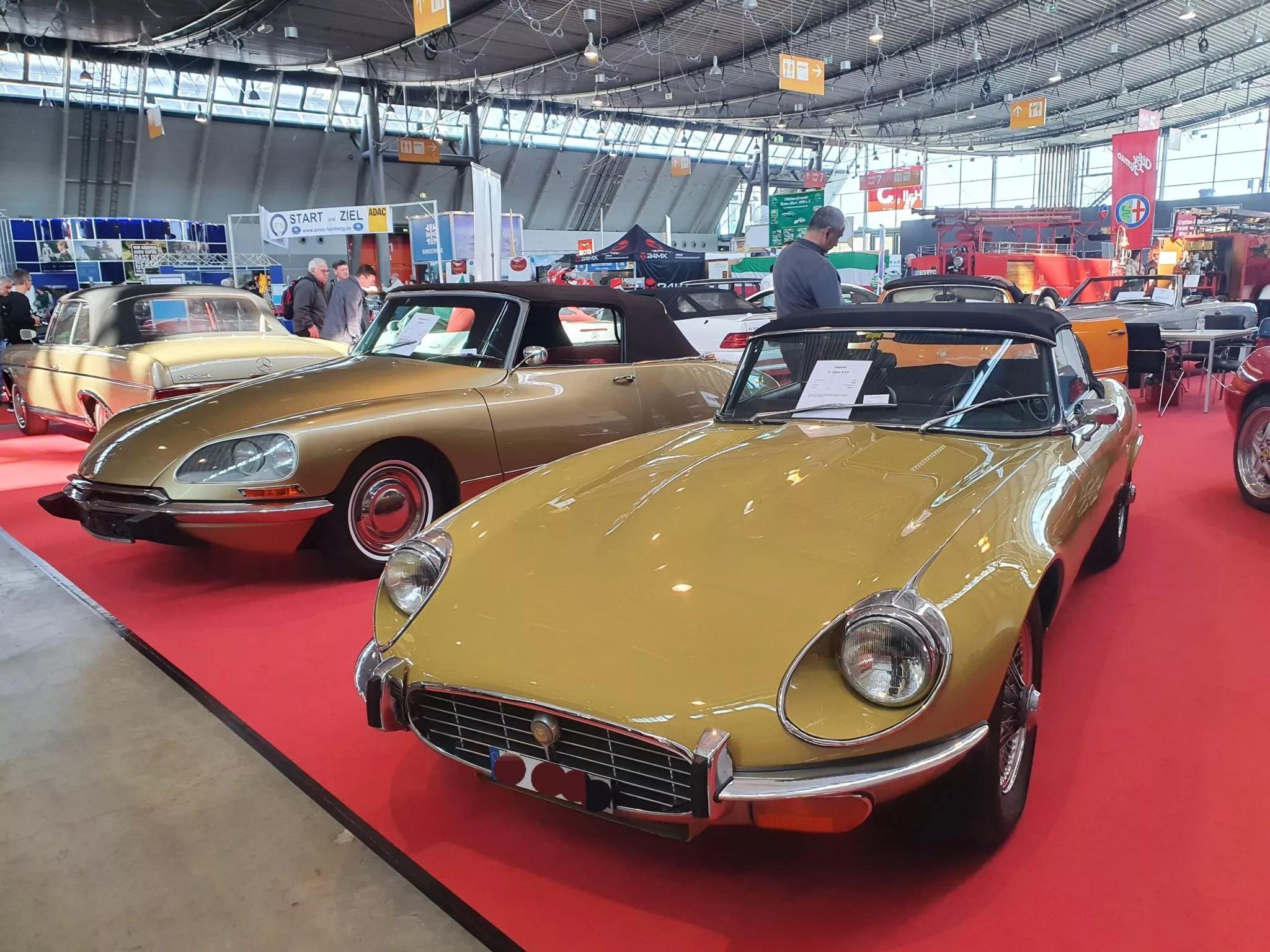 Zwei gelbe Oldtimer auf rotem Teppich bei einer Autoausstellung; vorne ein Cabrio mit schwarzem Verdeck und Schild an der Windschutzscheibe.