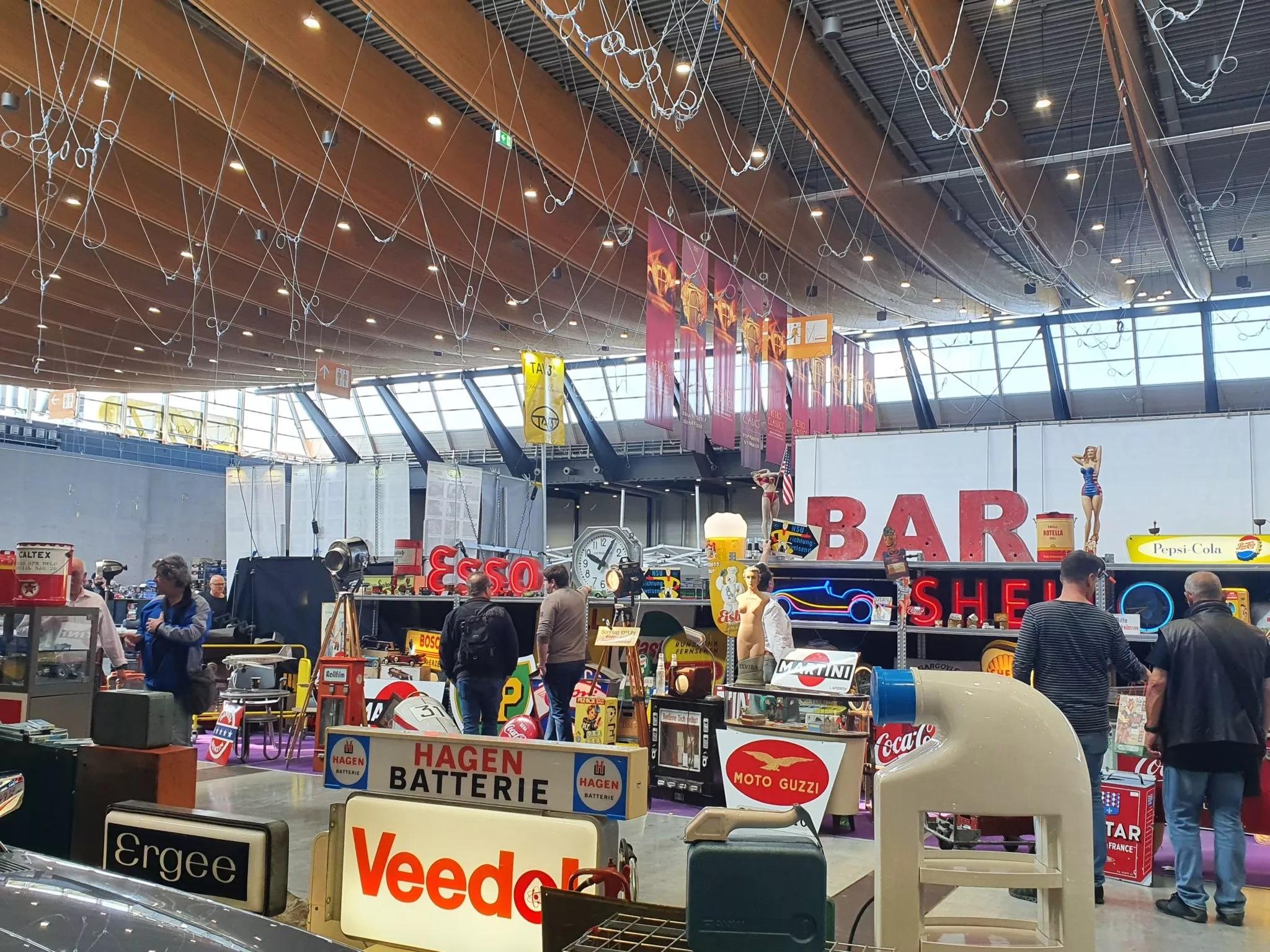 Hallenmarkt oder Ausstellung mit Vintage-Schildern wie ‚Esso‘, ‚SHELL‘ und ‚Moto Guzzi‘; Besucher schlendern zwischen den Exponaten.