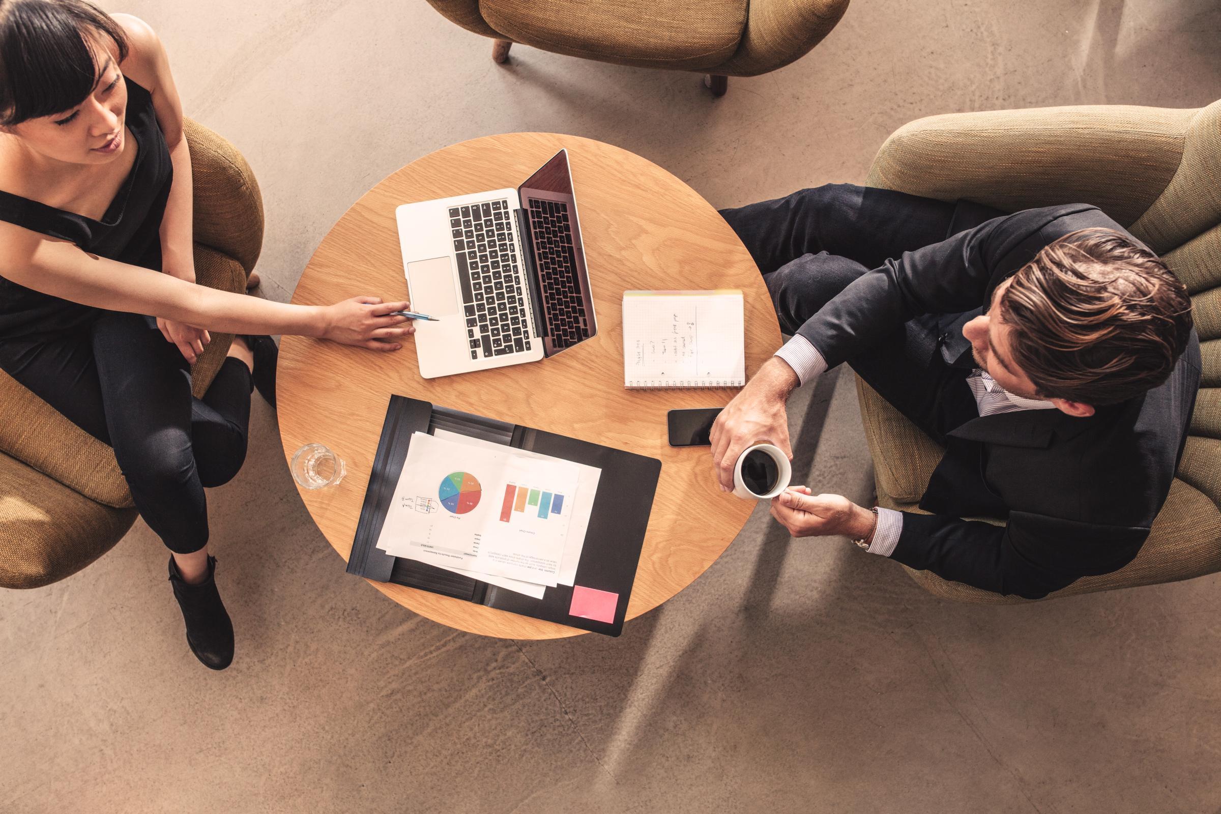 Zwei Personen sitzen an einem runden Holztisch. Auf dem Tisch liegen ein offener Laptop, ein Notizbuch mit handschriftlichen Notizen und eine Mappe mit Diagrammen und Grafiken. Eine Person zeigt mit einem Stift auf den Laptopbildschirm, die andere hält eine Tasse Kaffee.