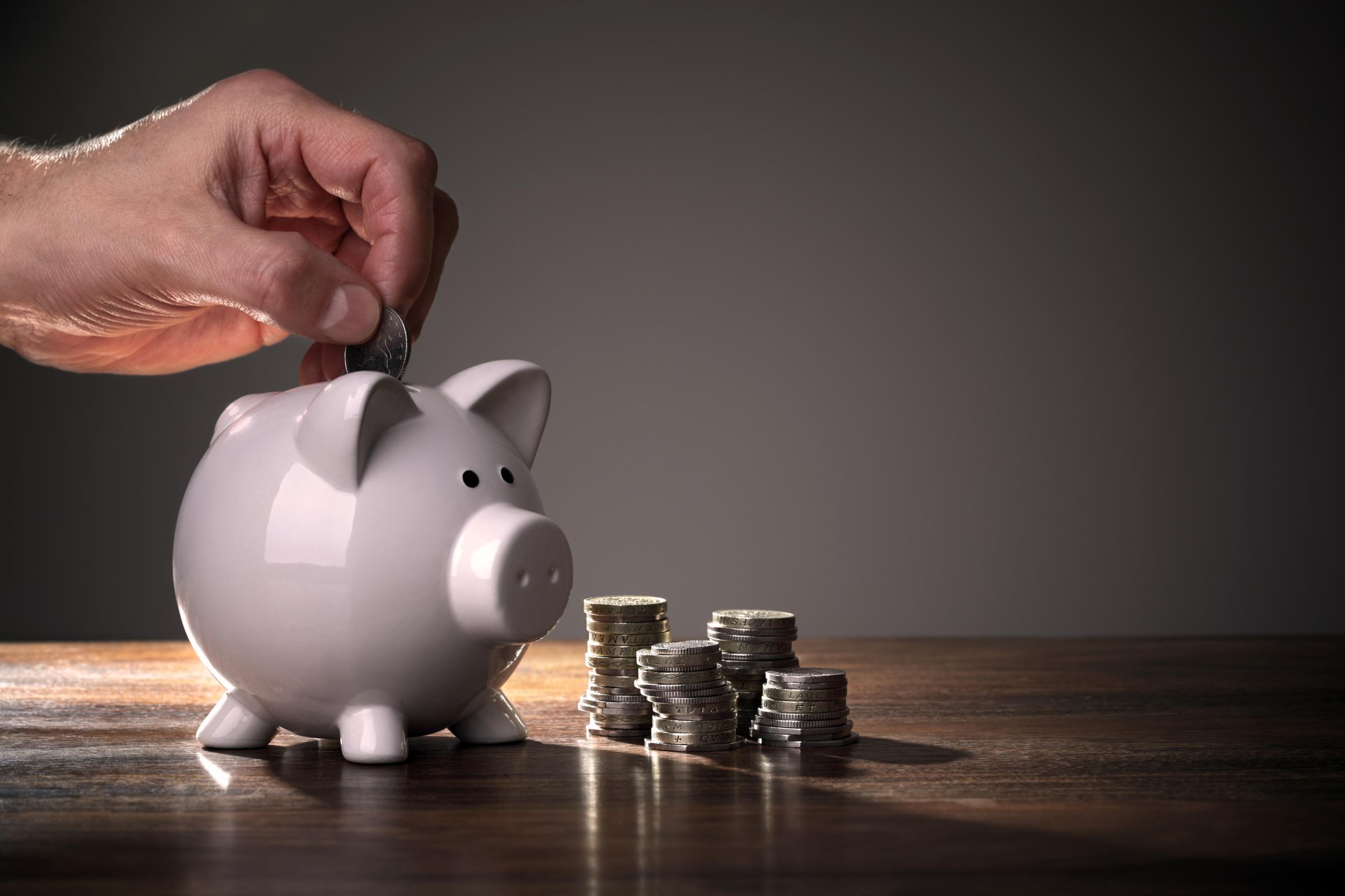 Eine Hand wirft eine Münze in ein weißes Sparschwein. Daneben stehen mehrere Münzstapel auf einer Holzoberfläche.