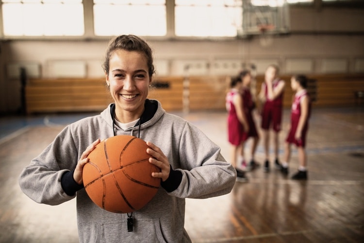 Person im grauen Hoodie hält einen Basketball in einer Sporthalle; im Hintergrund unterhalten sich mehrere Personen in roten Trikots.