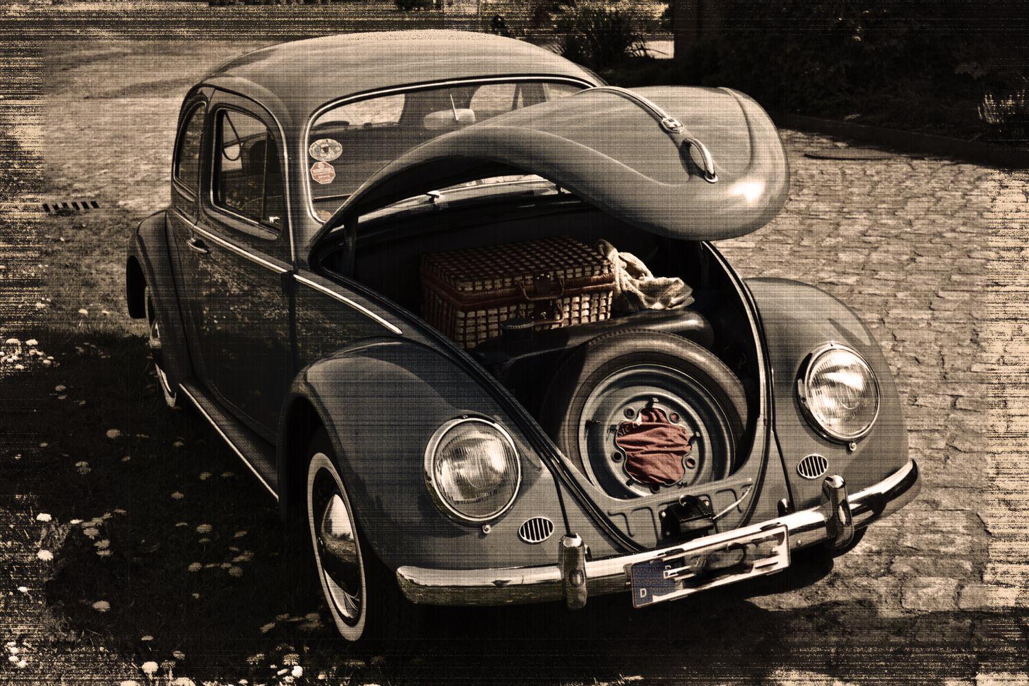 Ein klassischer VW Käfer mit geöffneter vorderer Haube, die einen Ersatzreifen, ein rotes Tuch und einen Picknickkorb im Kofferraum zeigt. Das Auto steht auf einem gepflasterten Weg, umgeben von Blumen.