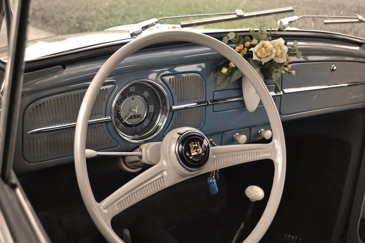 Innenraum eines Oldtimers mit weißem Lenkrad, blauem Armaturenbrett und einem kleinen Blumenstrauß auf dem Armaturenbrett. Ein blauer Schlüssel hängt am Lenkrad.