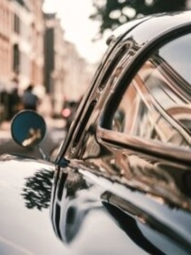 Ein schwarzer Youngtimer mit Fokus auf das Fenster und den linken Seitenspiegel. Im Hintergrund sind unscharfe Gebäude zu erkennen.