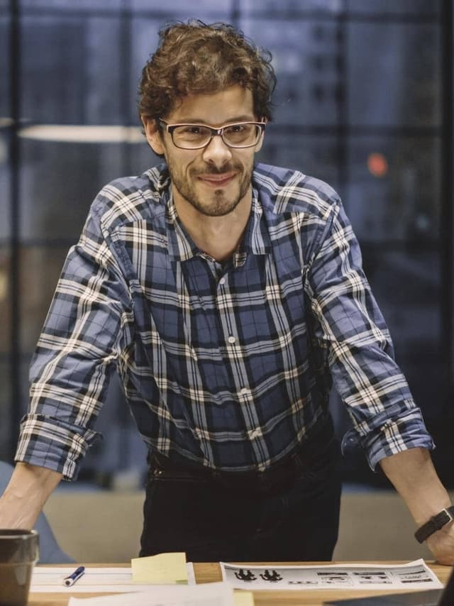 Ein Mann mit Brille stützt sich am Schreibtisch ab und lächelt direkt in die Kamera.
