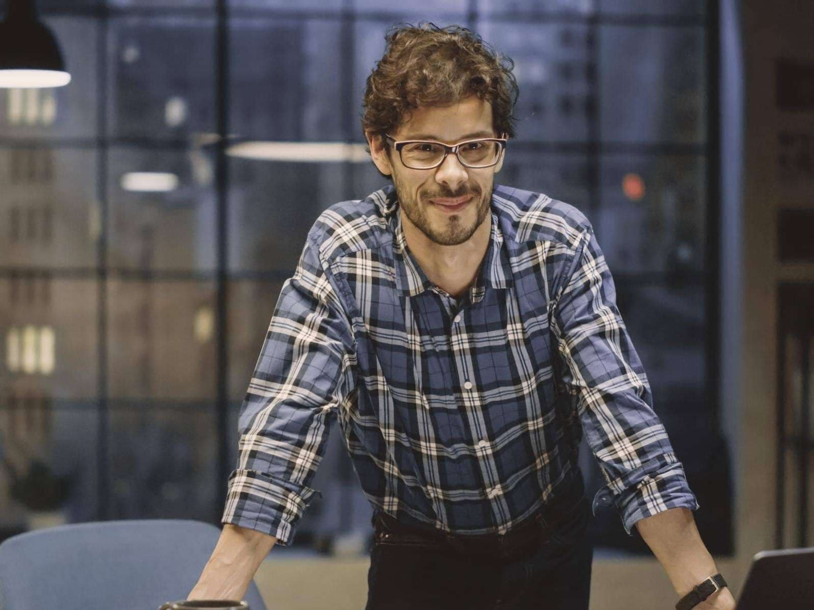 Ein Mann mit Brille stützt sich am Schreibtisch ab und lächelt direkt in die Kamera.