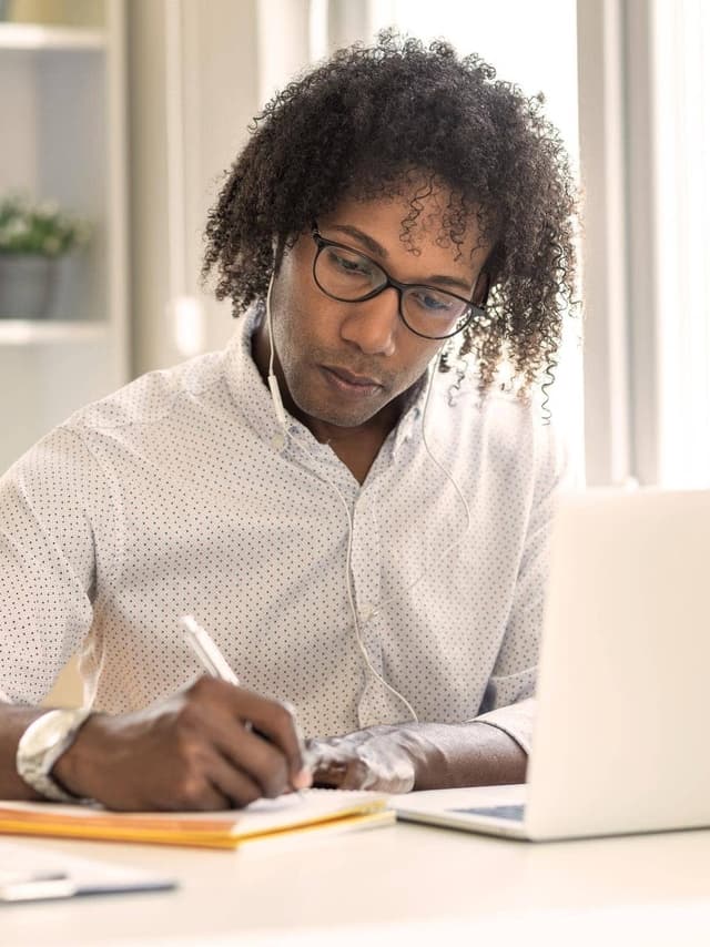 Ein Blogger mit Brille schreibt auf einem Blatt Papier, während er Kopfhörer trägt und an seinem Laptop arbeitet.