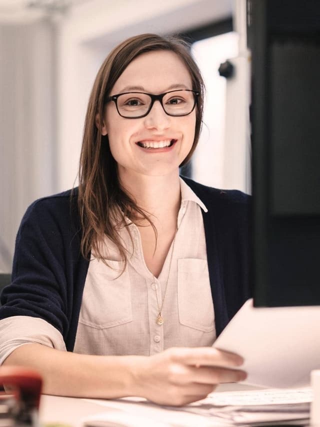 Eine Frau mit Brille sitzt an einem Schreibtisch mit einem Blatt Papier und lächelt.