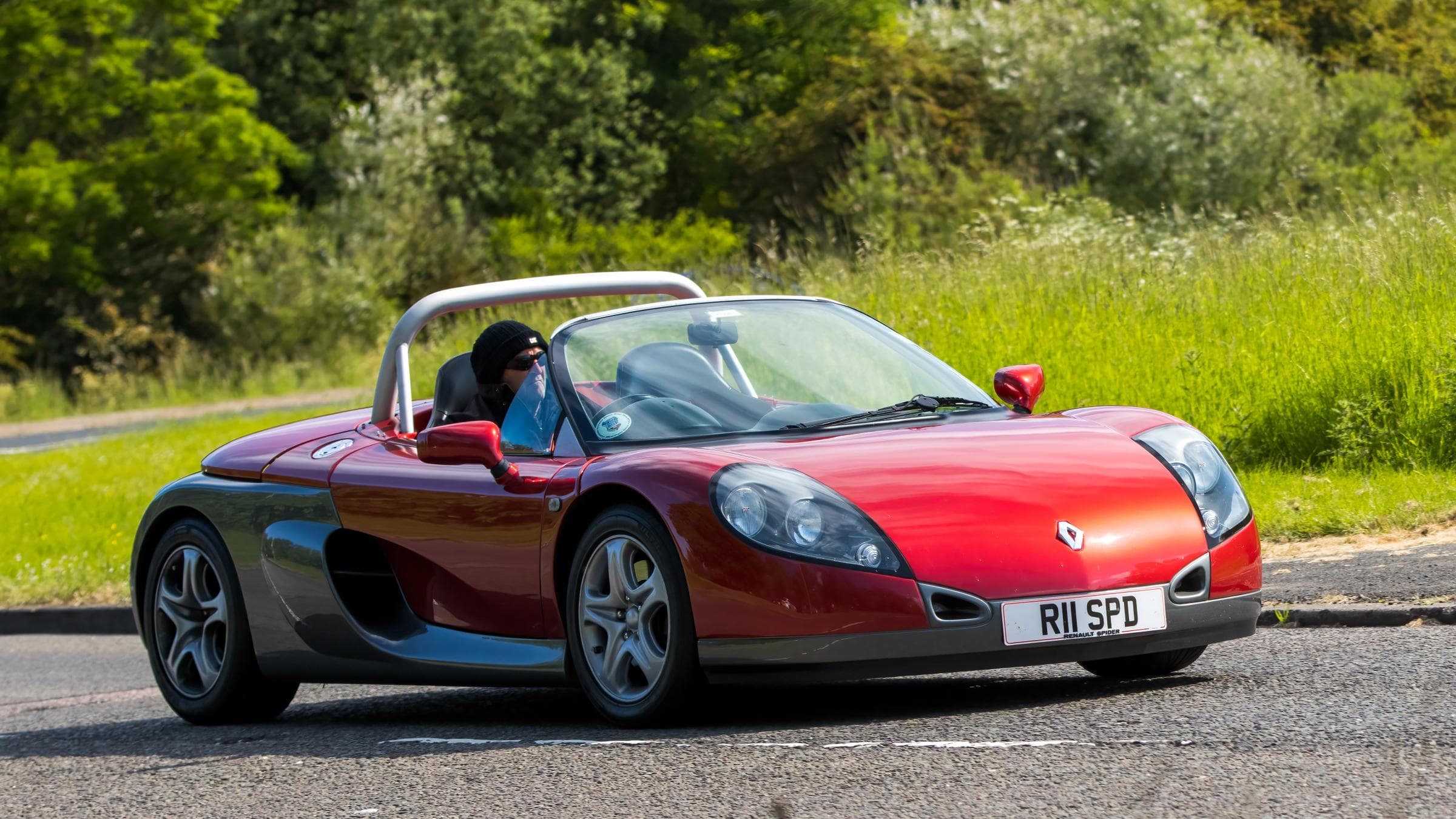 Roter Renault Sport Spider mit Fahrerin fährt auf einer geterrten Straße an einem sonnigen Tag.