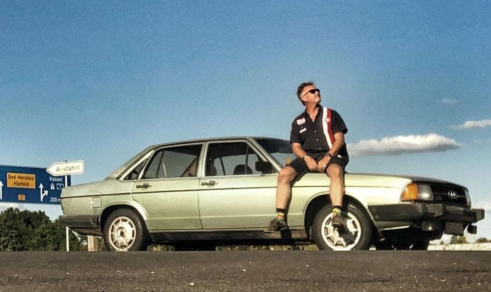 Ein Mann, Jenz Tanz, sitzt auf einem parkenden grünen Audi 100 Typ 43.