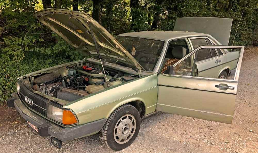 Ein Audi 100 Typ 43 steht am Waldrand geparkt. Der Motorraum, der Kofferraum und die Autotür sind geöffnet.