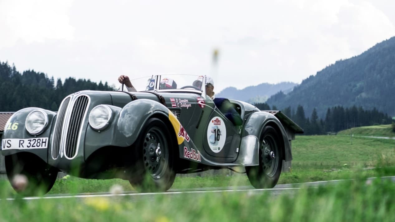 Dunkelgrüner Oldtimer fährt auf einer Landstraße mit Redbull Logo und zwei Fahrern mit weißen Caps. Im Hintergrund sieht man grüne Wiesen und eine Berglandschaft.