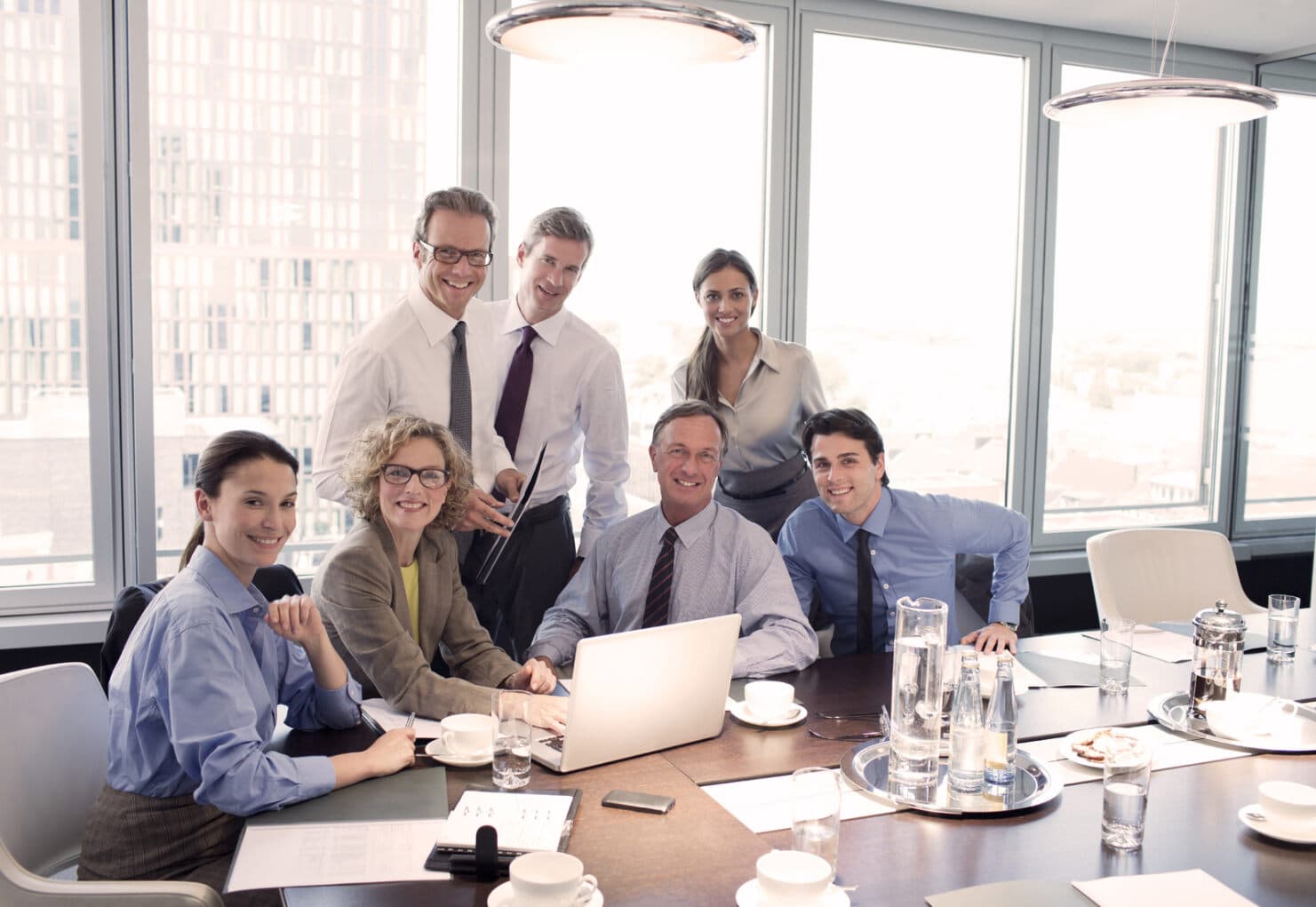 Sieben lächelnde Geschäftsleute sitzen um einen Laptop an einem Meetingtisch in Business-Kleidung.