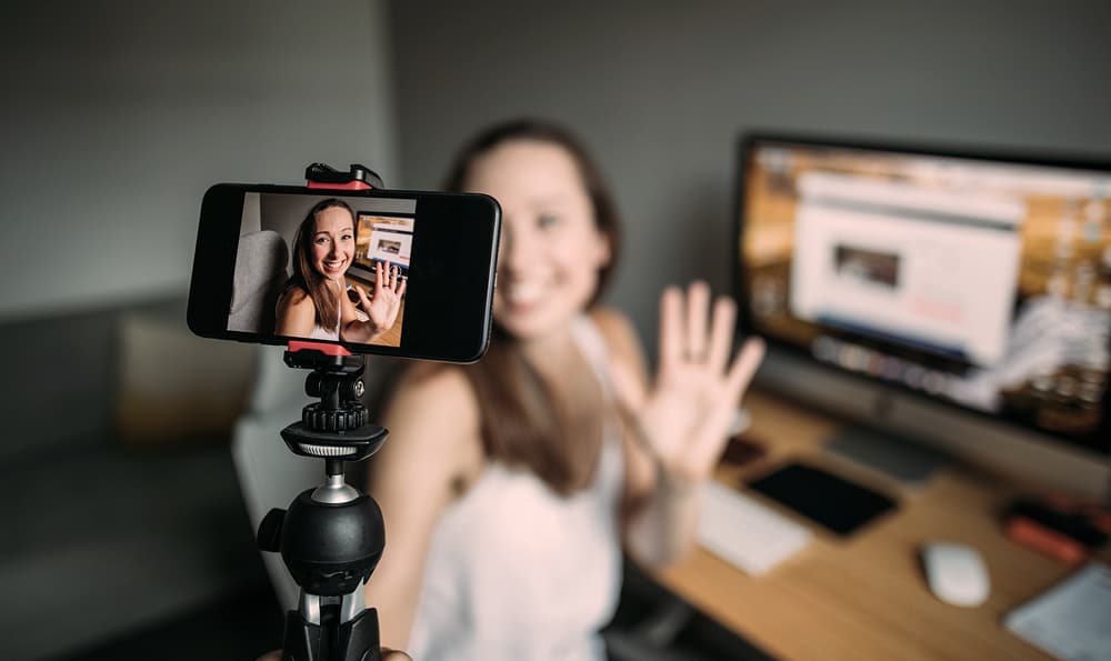Lächelnde Influencerin sitzt vor ihrem PC und hält eine Vlogging-Kamera in der Hand und filmt sich selber