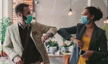 Zwei Personen tragen Gesichtsmasken in einem Café und begrüßen sich mit Ellbogen während der Corona-Pandemie.
