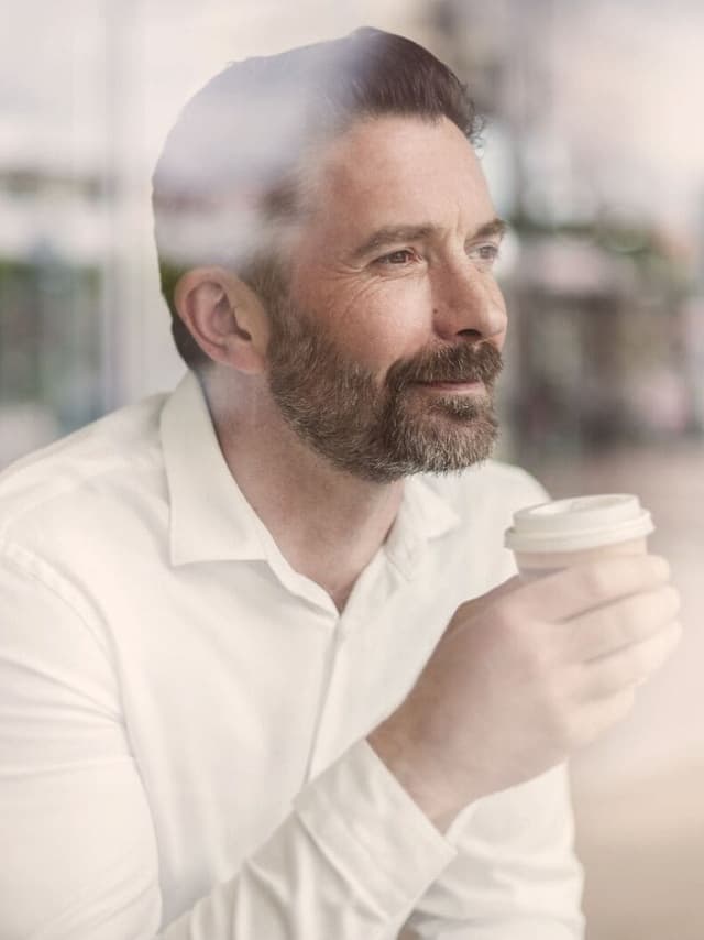 Ein Geschäftsmann hält eine Tasse Kaffee in der Hand und blickt aus dem Fenster eines Cafés in die Ferne.