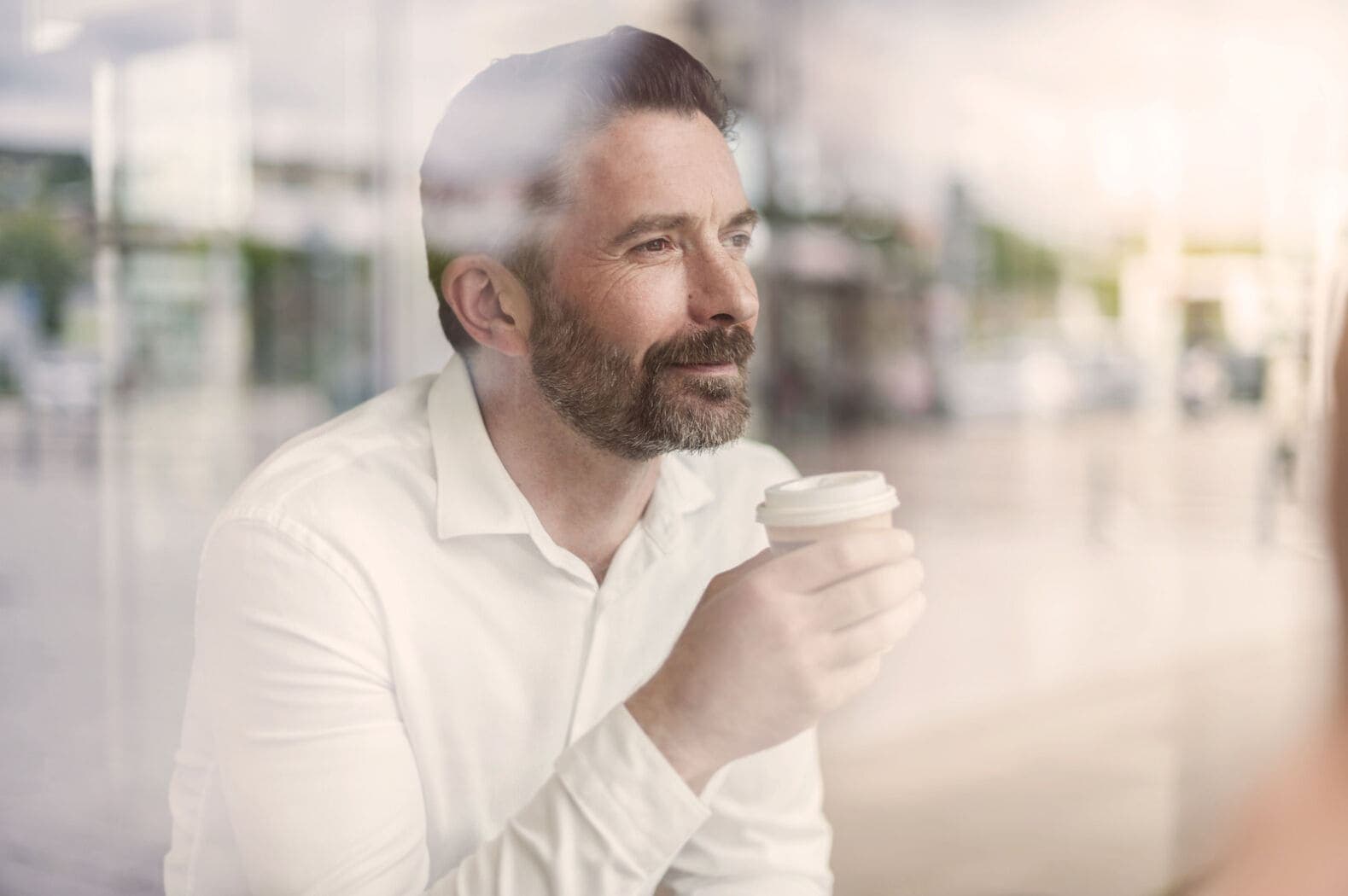 Ein Geschäftsmann hält eine Tasse Kaffee in der Hand und blickt aus dem Fenster eines Cafés in die Ferne.