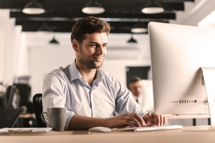 Junger Mann in Hemd und braunen Haaren sitzt lächelnd vor seinem PC. Im Hintergrund arbeitet ein weiterer Mann.
