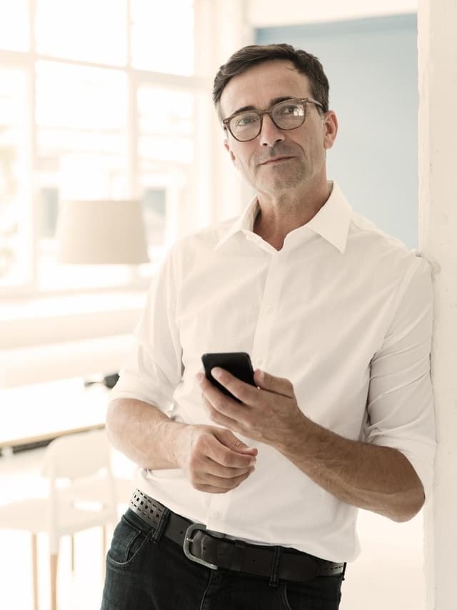 Personalberater-Versicherung von Hiscox: Headhunter in Hemd und Brille lehnt in der Tür seines Büros und hält ein Handy