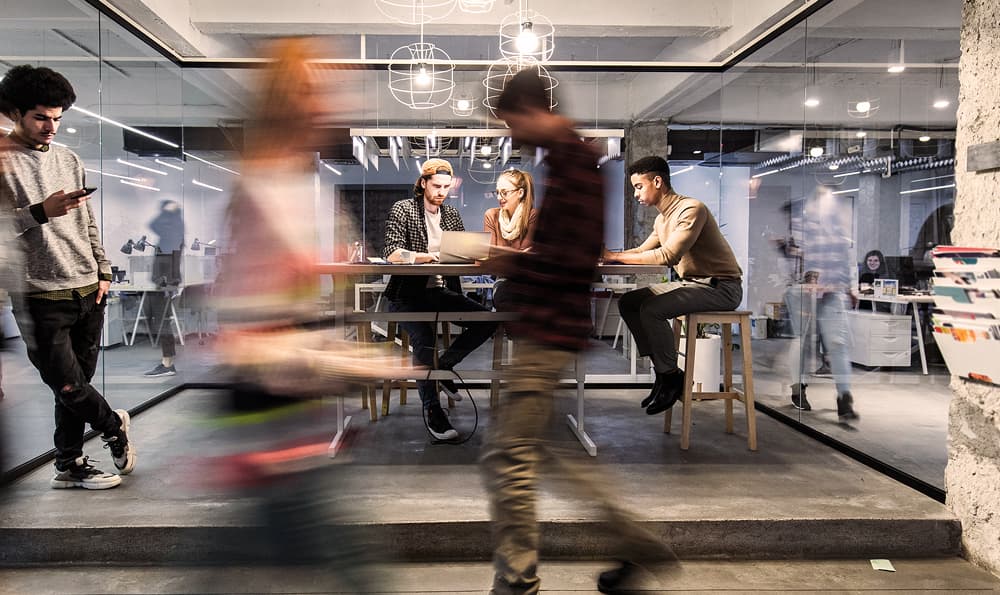 Ein volles Glasbüro eines Start-ups voll mit Mitarbeitern an ihren Laptops. Mitarbeiter laufen vorbei und gehen zu einem Meeting