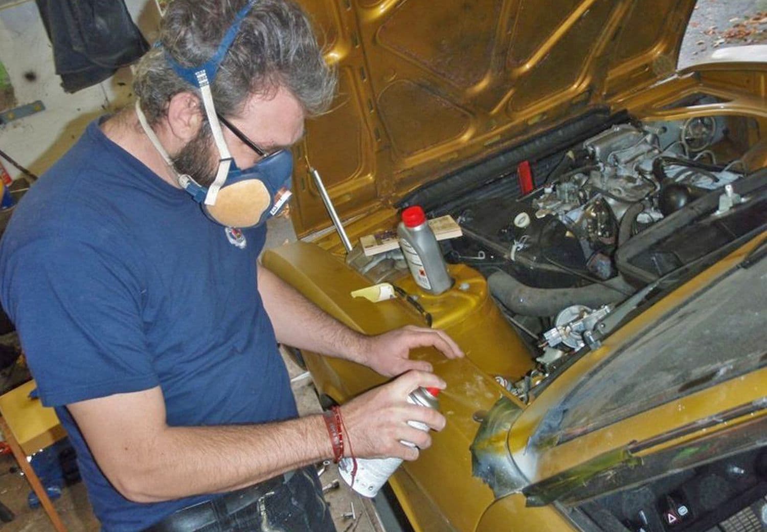 Wartungsarbeiten werden an dem Lack eines goldenen VW K70 L 1971 vorgenommen. Mann hält eine Lack-Sprühdose und trägt Atmenschutzmaske